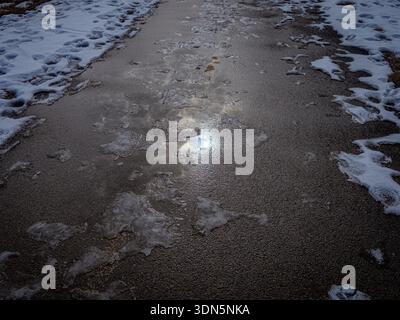 Partially melted ice and snow on asphalt path reflecting sunlight and clouds, capturing late winter thaw textures in soft natural light. Stock Photo