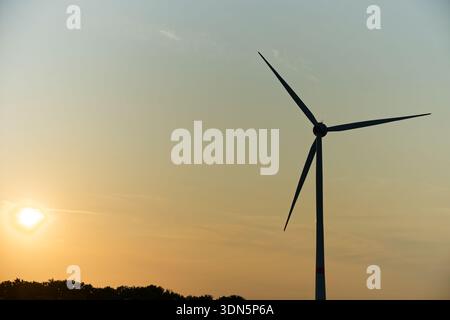 A single wind turbine stands alone on the horizon, its blades turning in dark silhouette while the sunset paints the sky in warm orange and gold tones Stock Photo