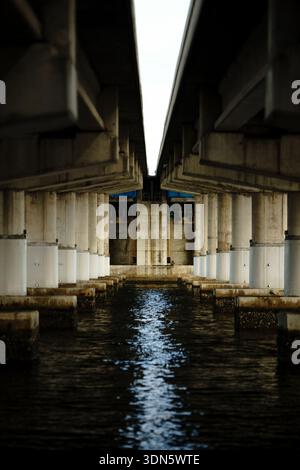 Symmetrical view under a concrete highway bridge with massive pillars reflecting in the water in Florida, USA. Industrial infrastructure. Stock Photo