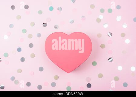 Red heart-shaper gift box on pink background with confetti. Valentines day concept. Top view, flat lay, mockup. Stock Photo