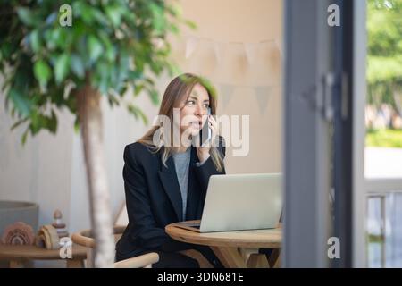 Confident businesswoman having phone conversation while working on remote in stylish city cafe. Stock Photo
