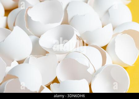 Small pieces of white eggshell are scattered on a bright yellow surface. The eggshells are from eggs that have been used for cooking or baking. Stock Photo