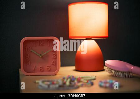 Still life showing pink alarm clock, table lamp with orange shade, hairbrush and colorful beaded jewelry arranged on wooden surface, objects illuminated by warm light Stock Photo