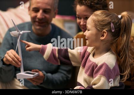 Caucasian middle aged man and woman sitting with Caucasian child girl, smiling and interacting with wind turbine model, child girl pointing at model while adults watching Stock Photo