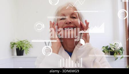 Smiling senior woman touching face in bathroom, wearing white bathrobe with AR overlays and plants Stock Photo