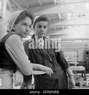 Sloviansk, Donetsk region, Ukraine, USSR. 1960s. Archival historical photograph. Two young female workers standing together during a break at the Sloviansk Ceramic Combine. The woman on the left wears a vest and a light headscarf, while her colleague wears a dark protective work coat. They lean on a structure, looking sideways with a calm expression. The background shows the blurred interior of the spacious workshop. This authentic black and white image captures the friendship and daily life of Soviet youth in the peaceful Donbas region Stock Photo