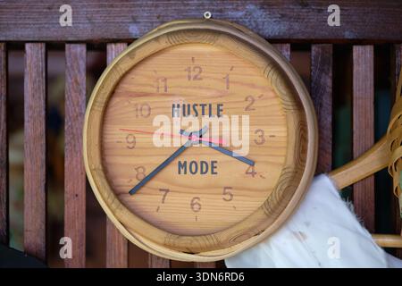 A natural wood wall clock displaying HUSTLE MODE on its face, with black and red hands. It hangs on a rustic slatted wooden wall inside a building Stock Photo