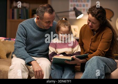 Caucasian middle aged man and Caucasian middle aged woman sitting with Caucasian child reading book together, child smiling while adults engaging and supporting learning activity Stock Photo