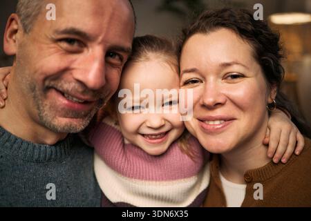 Portrait of Caucasian middle aged man, Caucasian woman, and Caucasian child girl smiling closely together, child girl hugging adults, all looking into camera Stock Photo