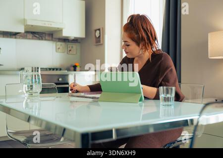 A young woman with red hair sits at a modern kitchen table, diligently writing in her journal while using a digital tablet and smartphone for referenc Stock Photo