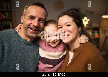 Portrait of Caucasian middle aged man, woman, and Caucasian child smiling together, child hugging adults, all looking into camera, close family moment Stock Photo