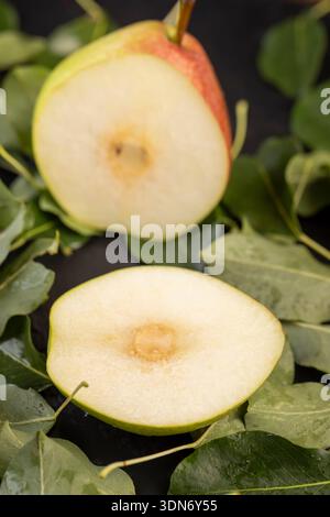 a ripe juicy sliced pear lies on the table with the green foliage of the pear, a large number of green leaves of the pear and a ripe covered pear with Stock Photo