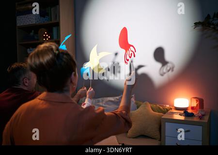 Middle aged Caucasian woman and middle aged Caucasian man with child creating animal shadow puppets on wall using colorful paper cutouts, sitting together in cozy bedroom setting with soft lighting Stock Photo
