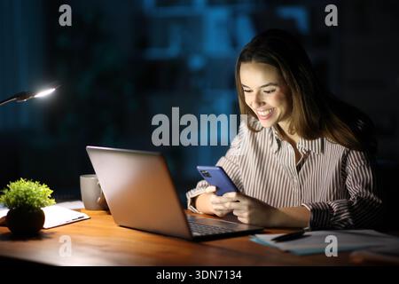 Happy tele worker in the night working using phone and laptop at home Stock Photo