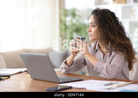 Serious freelancer holding coffee looking away sitting at workplace at home Stock Photo
