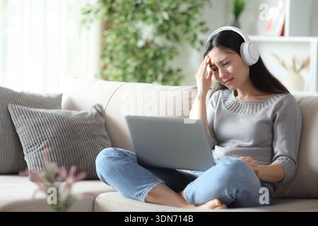Worried asian woman wearing headphone is checking laptop sitting on a sofa at home Stock Photo