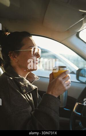 Woman sits in a car holding a travel mug, smiling and enjoying the moment. Sunlight streams through the window, creating a warm and relaxed atmosphere Stock Photo