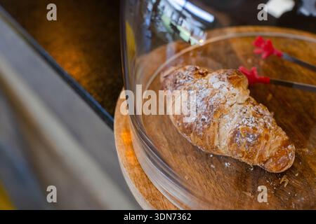 Single croissant resting on round wooden board under dome cover with metal tongs on bakery counter Stock Photo