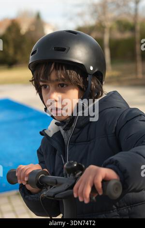 Child wearing winter jacket and helmet, holding scooter handles, looking ahead in a park Stock Photo