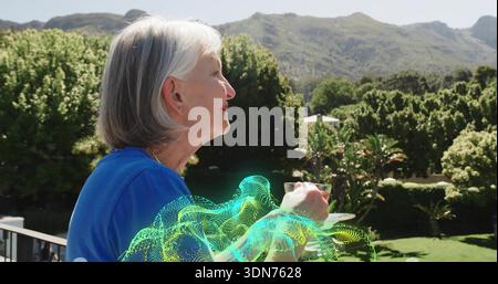 Standing senior woman holding glass cup saucer on balcony in bright blue top, hologram overlay Stock Photo