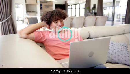 Reclining adult man in coral t-shirt browsing silver laptop on sectional couch, curtains visible Stock Photo