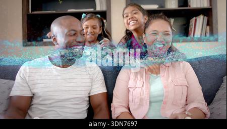 Laughing family of four sitting on gray couch at home, two girls leaning, teal-blue band crossing Stock Photo