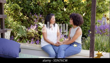 Holding hands, mother and daughter in sleeveless tops on garden wall under pergola, with gold stars Stock Photo