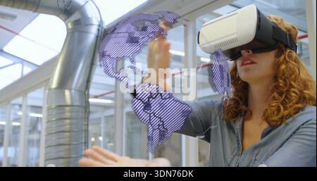 Reaching woman wearing white VR headset, denim shirt, manipulating purple hologram map in tech lab Stock Photo