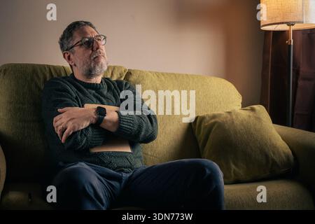A mature man with glasses and a beard sits on a green sofa at home, looking up thoughtfully while holding a folder, representing senior mental wellbei Stock Photo