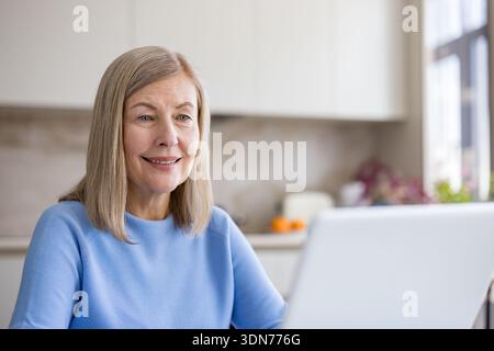 Senior woman at home smiling while having a video call or working remotely on her laptop in a modern kitchen, representing active aging and digital co Stock Photo