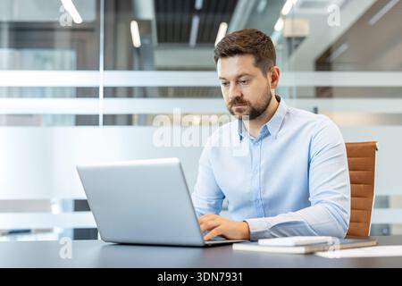 Concentrating man with beard using a laptop computer at a desk in a contemporary corporate workspace, actively engaged in freelance work or business tasks Stock Photo