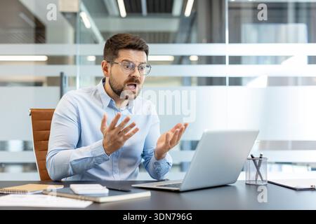 Young man wearing eyeglasses feeling stressed and angry, actively gesturing while having a difficult discussion or conflict during a virtual meeting on his laptop in a modern workplace Stock Photo