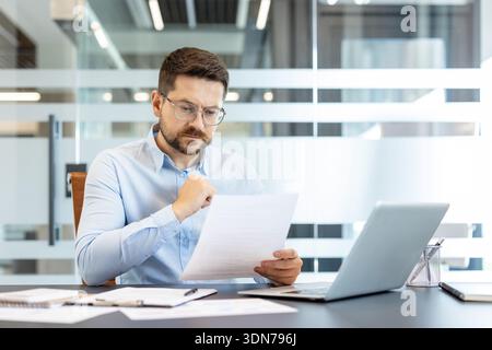 Businessman wearing glasses and a light blue shirt. Sitting at a desk with a laptop. Concentrating on reading documents and analyzing important corporate paperwork in a contemporary office environment Stock Photo