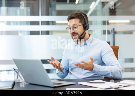 Professional man wearing a headset and glasses, actively gesticulating during a video call on his laptop, working remotely from a contemporary office environment Stock Photo