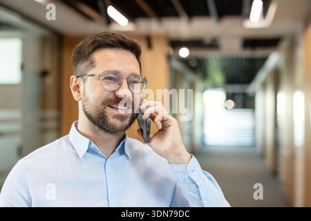 Professional man happily engaging in a mobile phone conversation, connecting with colleagues and networking while standing in a busy corporate hallway Stock Photo