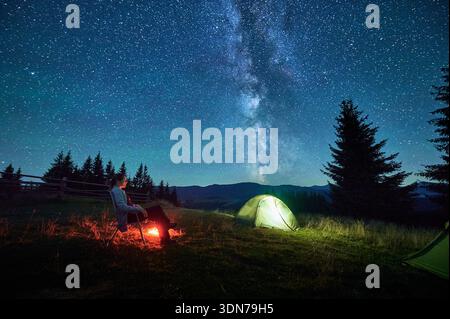 Hiker woman relaxes by campfire, illuminated by its warm glow, with tent nearby under stunning, star-filled sky. Milky Way stretches overhead, adding magical touch to serene mountain setting. Stock Photo