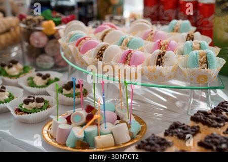 A vibrant assortment of pastel-colored macarons, marshmallows, and assorted desserts arranged on tiered glass stands at a festive party setting with b Stock Photo