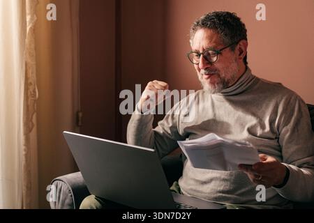 Relieved middle aged man smiling while reading documents on a laptop at home, feeling satisfied after solving a financial problem and completing paper Stock Photo