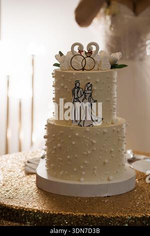 A beautifully decorated two-tier wedding cake with white frosting, pearl embellishments, and a swan ring topper, set on a shimmering gold tablecloth d Stock Photo