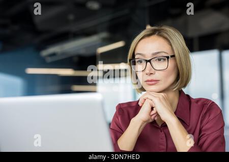 Businesswoman with glasses concentrating on her laptop screen, forming hypotheses, making plans, and seeking solutions while working in an open office environment Stock Photo