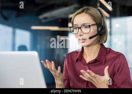 Stressed customer service representative wearing a headset, communicating with clients and indicating confusion or problems while working on a laptop in a modern office Stock Photo