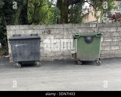 Two trash bins, one black and one green, are located next to a stone wall. The bins stand on a road with trees in the background. The setting is durin Stock Photo