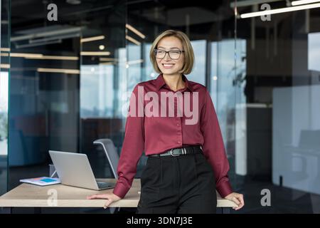 Young businesswoman standing in a contemporary office, smiling confidently and looking away, showcasing professionalism, leadership, and success in her career Stock Photo