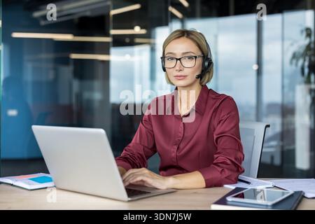 Focused woman wearing a headset and glasses, sitting at an office desk, actively working on a laptop, providing professional customer service and technical support Stock Photo