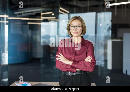 Professional female executive with arms crossed, looking confidently at the camera in a modern glass-walled office, conveying leadership, ambition and corporate success Stock Photo