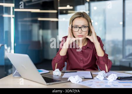 Young businesswoman with a serious expression and hands on her temples, feeling overwhelmed and frustrated at her desk with crumpled papers and a laptop, indicating stress and a challenging workload Stock Photo