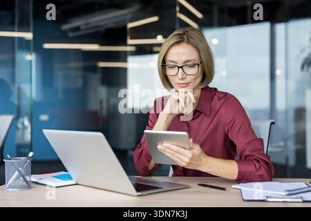 Professional businesswoman wearing eyeglasses is concentrating while using a digital tablet at her desk, with a laptop and documents nearby, embodying modern work and technology integration Stock Photo