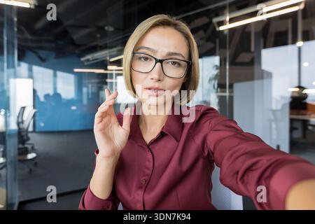 Young businesswoman wearing glasses having online video conference, discussing business at remote work, communicating with colleagues or clients via webcam in contemporary workspace Stock Photo