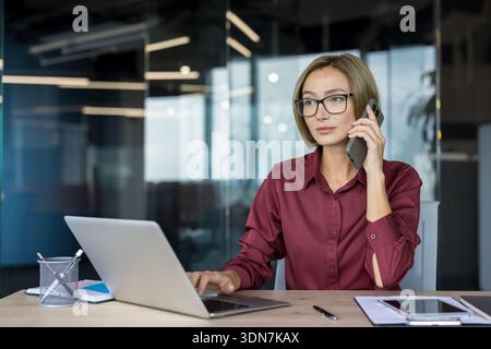 Confident businesswoman talking on a smartphone while simultaneously typing on a laptop, demonstrating multitasking and efficient communication in a professional office environment Stock Photo