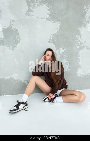 Slender woman posing in a trendy brown sweatshirt and white shorts. Sitting cross-legged on the floor against a textured plaster background. Natural l Stock Photo